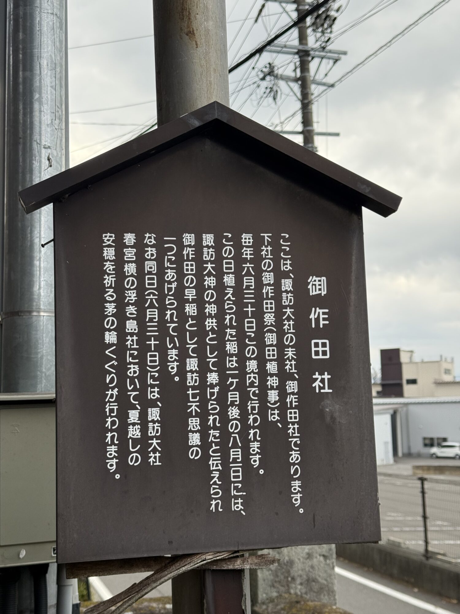 長野県下諏訪にある御作田社の木製の由緒書き看板。諏訪大社の末社であることや、毎年6月30日に行われる御作田祭（御田植神事）、そして植えてから1ヶ月で収穫し神供として捧げられたという「諏訪七不思議」の一つ「御作田の早稲」についての説明文が記載されている。