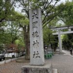 東京都府中市にある大國魂神社の入り口の風景。「大國魂神社」と大きく彫られた立派な社号標（石柱）が手前に建っており、その奥には大きな石造りの鳥居と、新緑の木々が連なる清々しい参道が続いている。