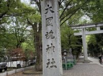 東京都府中市にある大國魂神社の入り口の風景。「大國魂神社」と大きく彫られた立派な社号標（石柱）が手前に建っており、その奥には大きな石造りの鳥居と、新緑の木々が連なる清々しい参道が続いている。