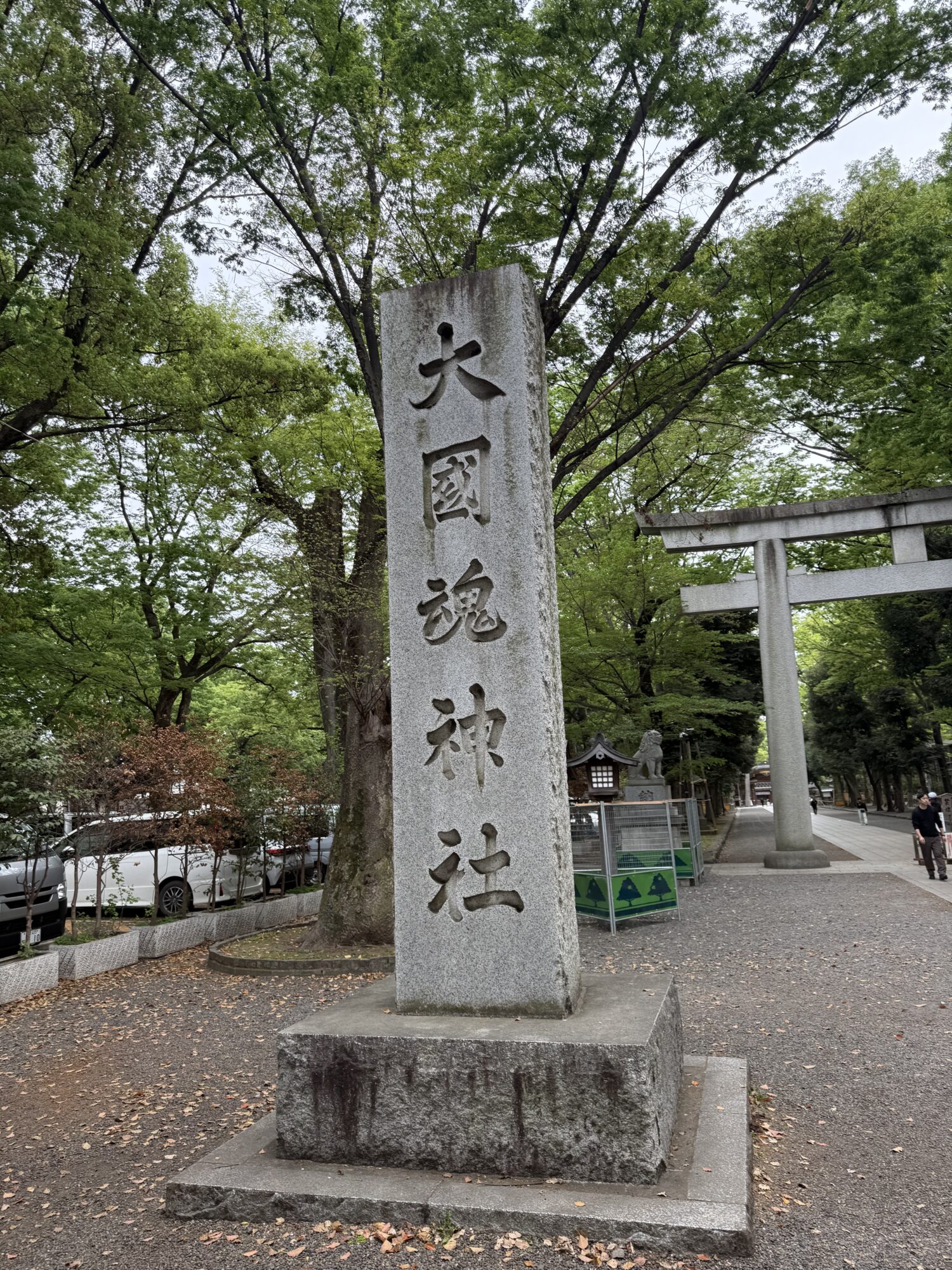 東京都府中市にある大國魂神社の入り口の風景。「大國魂神社」と大きく彫られた立派な社号標(石柱)が手前に建っており、その奥には大きな石造りの鳥居と、新緑の木々が連なる清々しい参道が続いている。