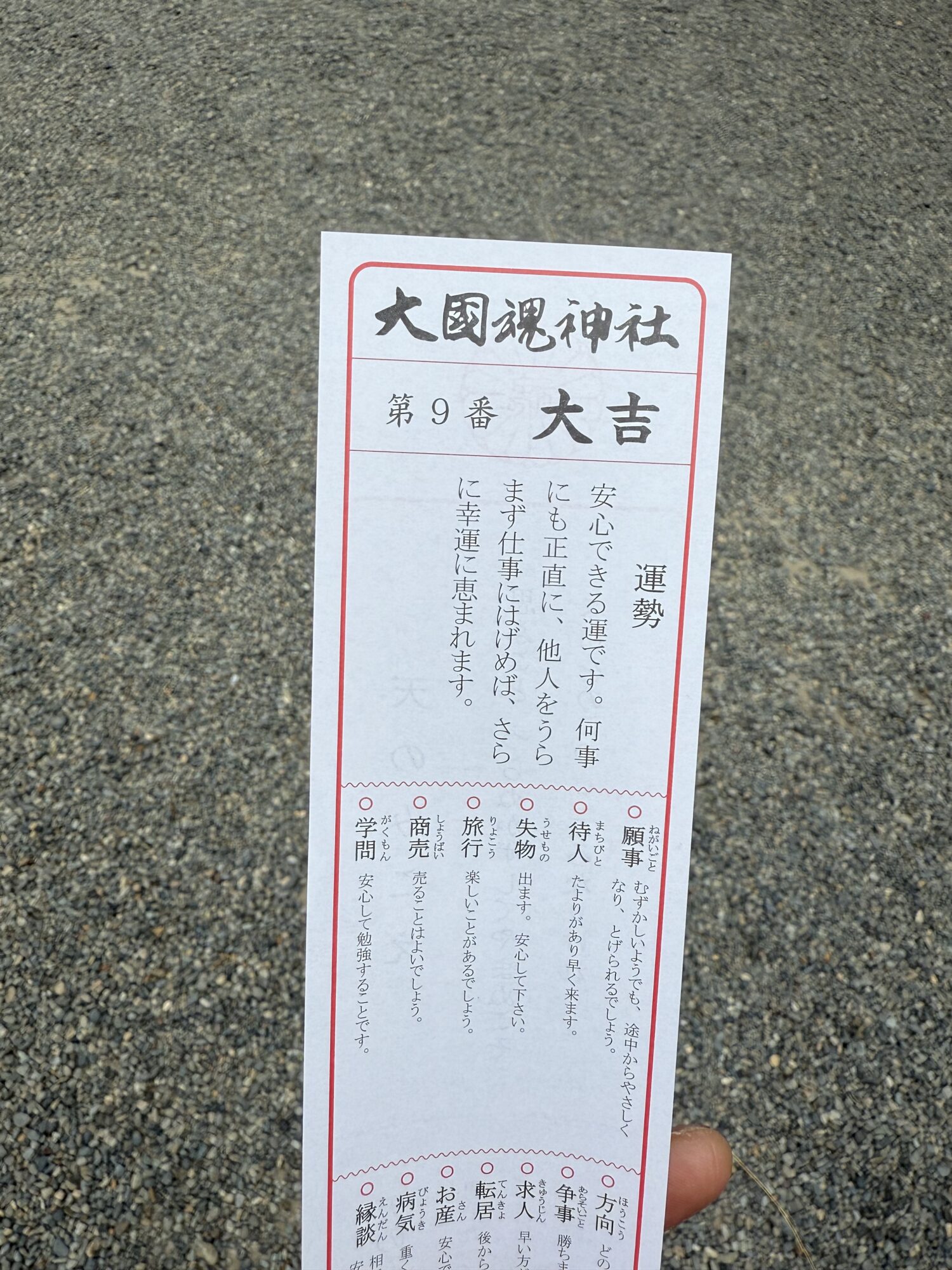 大國魂神社で引いた第9番のおみくじの写真。結果は最も良い「大吉」。「安心できる運です。何事にも正直に、他人をうらまず仕事にはげめば、さらに幸運に恵まれます」という運勢の文章がはっきりと読み取れる。背景には境内の玉砂利が写っている。