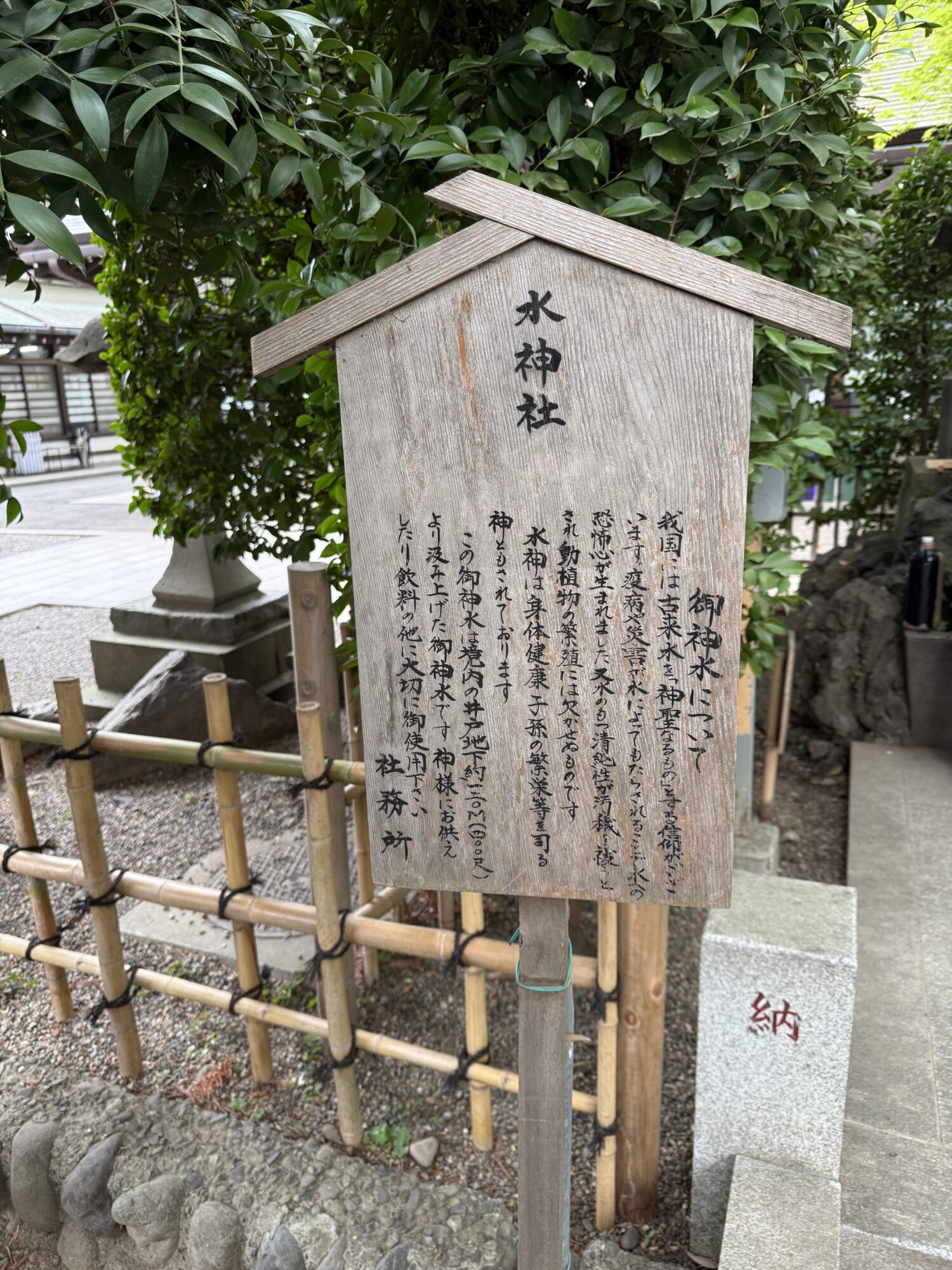 大國魂神社の拝殿左脇にある「水神社」の木製立て札の写真。立て札には「御神水について」として、水神が身体健康や子孫繁栄等を司ること、そしてこの水が境内地下約100メートルから汲み上げられた神聖な御神水である旨が墨文字で記されている。