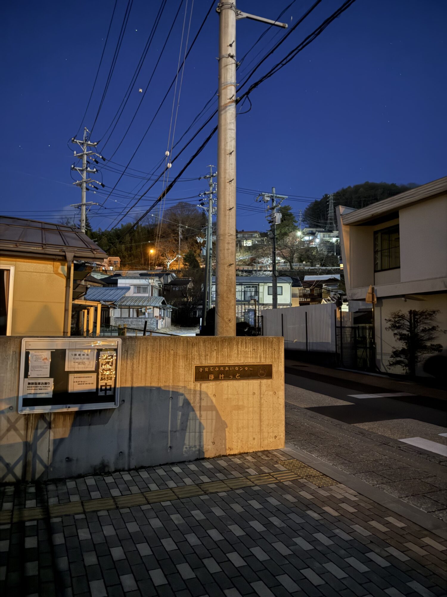 お水取りの吉時刻を待機している諏訪市周辺の夜の風景。17時半頃の撮影。高画質カメラの補正により夜空が青く明るく写っているが、実際は真っ暗。手前にはコンクリートの壁と電柱があり、奥には街灯に照らされた坂道と住宅街が見える。