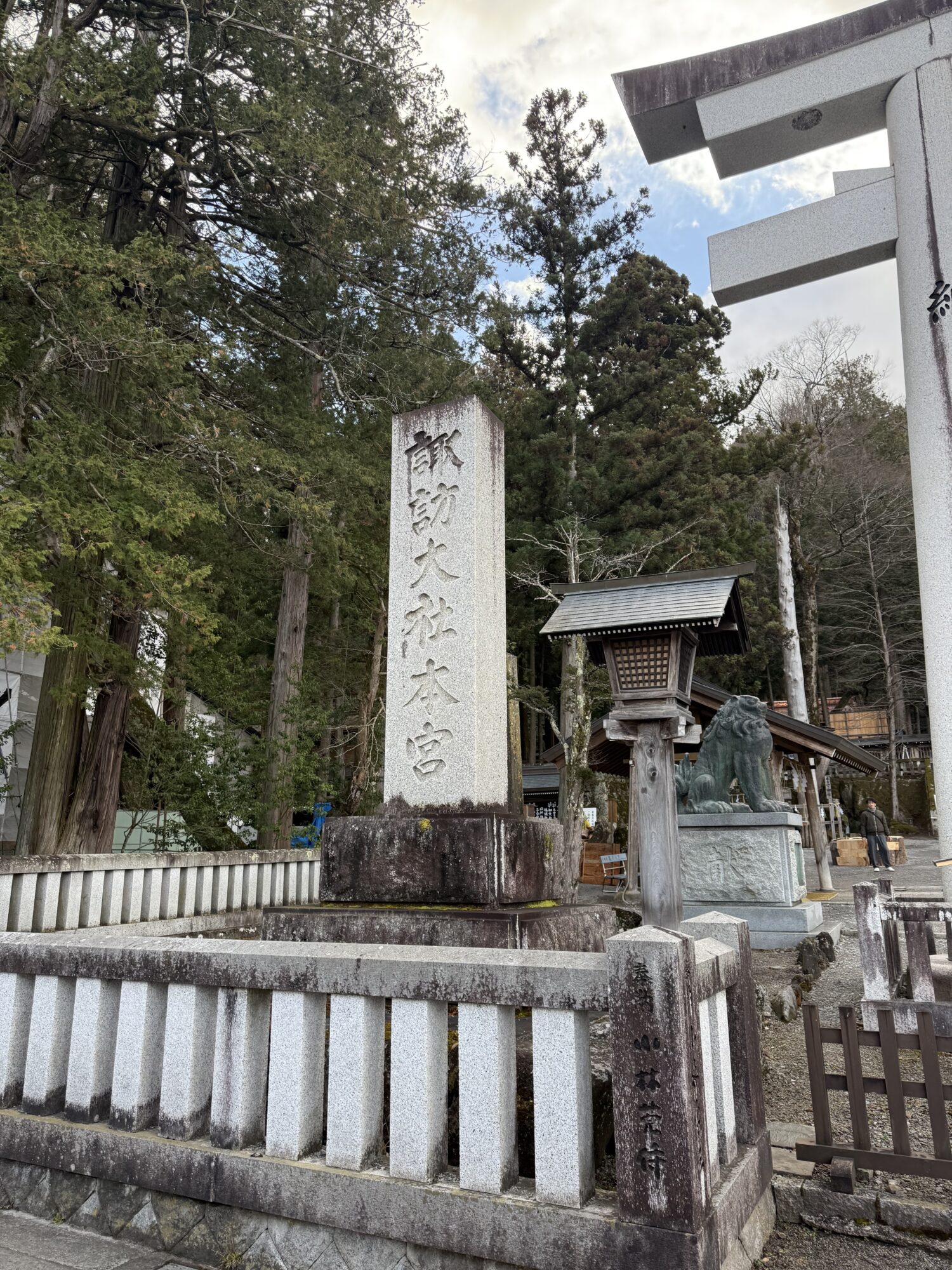長野県にある諏訪大社上社本宮の境内の風景。「諏訪大社本宮」と深く刻まれた立派な石柱（社号標）を中心に、右側には白い大鳥居の一部や石灯籠、狛犬が配置されている。背景には高くそびえる木々が茂り、昇殿参拝後の清々しい空気が感じられる。